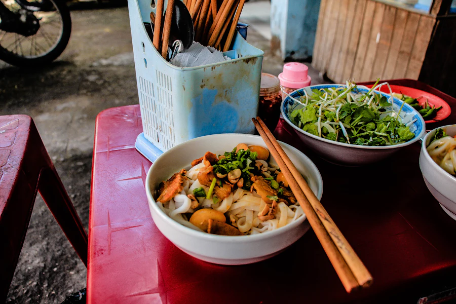 A bowl of noodle soup with fresh herbs and chopsticks on a street-food table